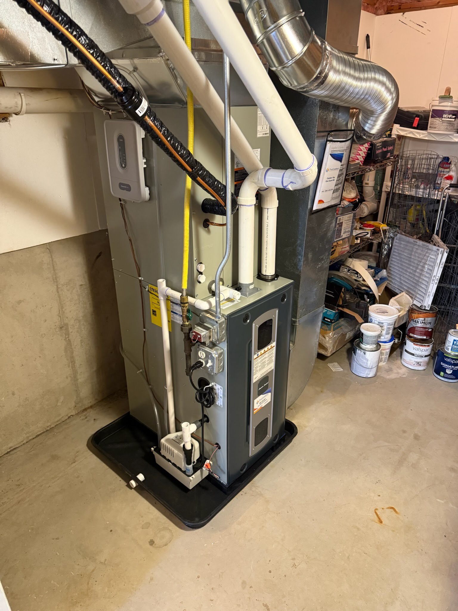 A home heating and cooling system (HVAC unit) installed in a basement, with visible ductwork, PVC pipes, electrical components, and a condensate pump, positioned on a plastic drip tray. The basement also has concrete flooring and various household items stored nearby.